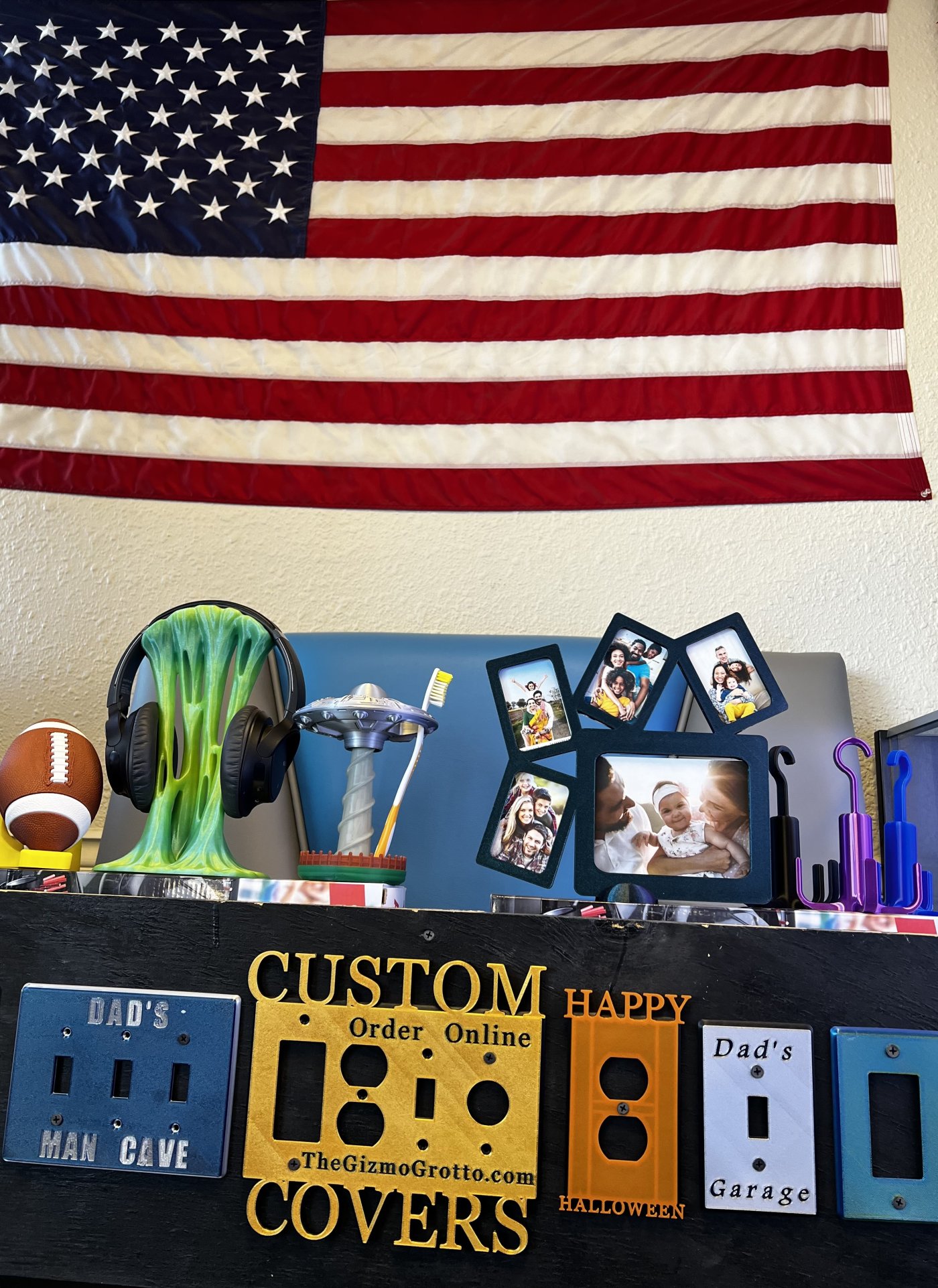 A shelf of custom 3D-printed products, including light switch covers, phone stands, and picture frames, under an American flag